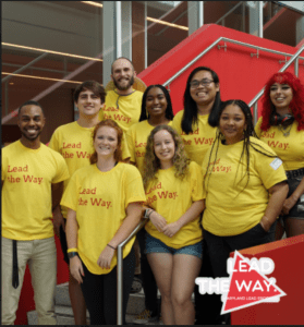 This is a photo of MLEAD facilitators. They are all in yellow t-shirts that say, "Lead the Way" in red. They are standing on the stairs in the Edward St. John Learning & Teaching Center.